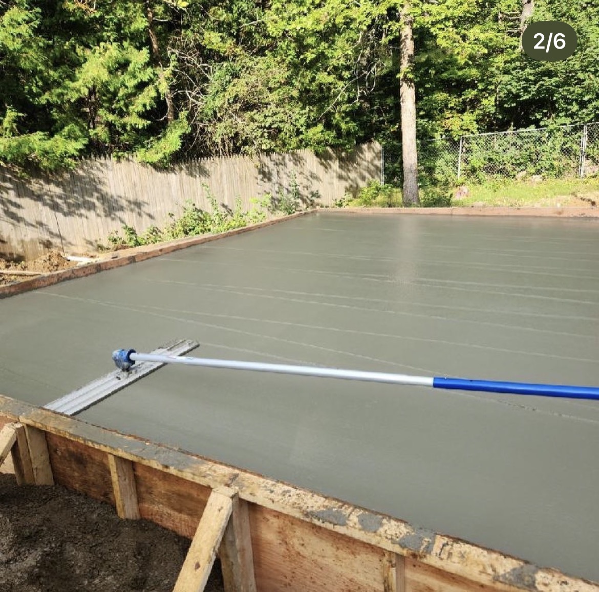 Freshly floated concrete slab inside wood forms beside a wooded backyard